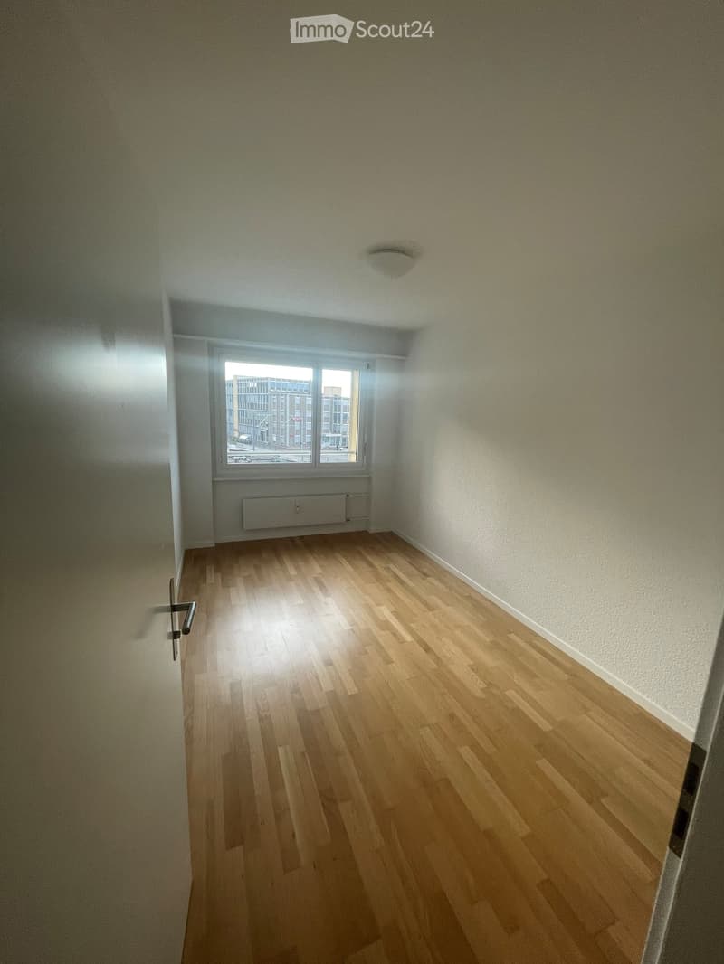 Empty room with wooden flooring, white walls, and a window showing a cityscape view. No furniture or equipment present.