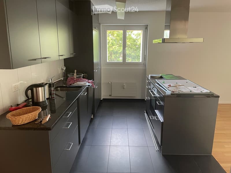 Modern kitchen with dark grey cabinets, black countertops, and stainless steel appliances. Features a central island, sink, and various kitchen items including a coffee maker and wicker basket.