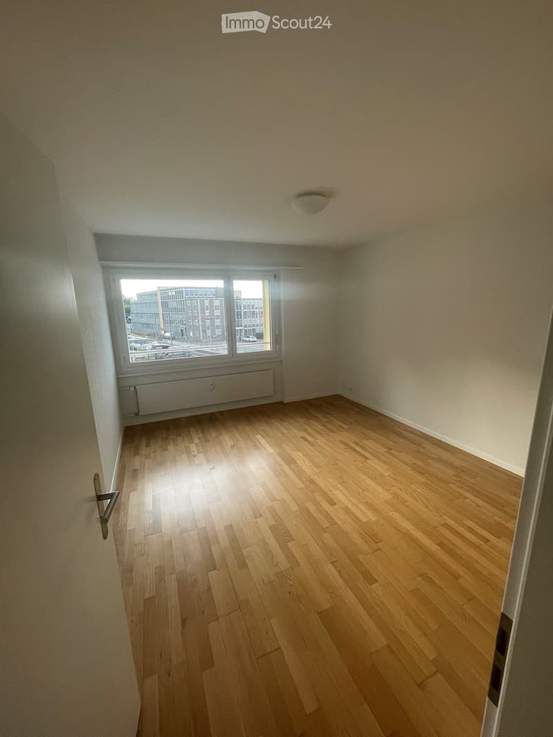 Empty room with wooden flooring, white walls, and large windows offering a view of the outside buildings.