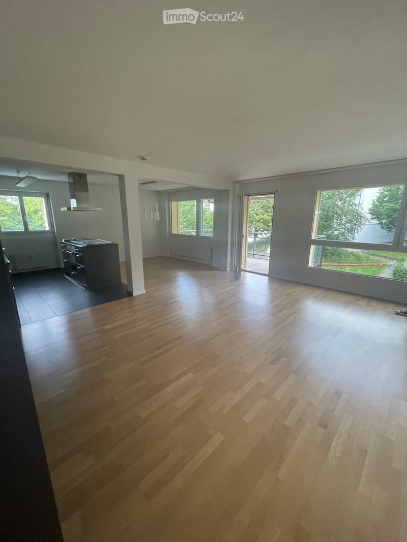 Empty room with wooden flooring, large windows, and a kitchen area visible in the background.
