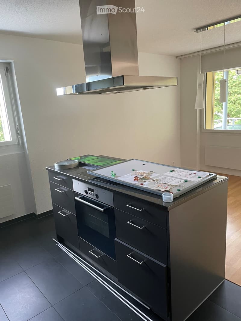Modern kitchen with dark grey cabinets, a central island, built-in oven, and a stainless steel range hood. The countertop holds various items including papers, a scale, and small containers.