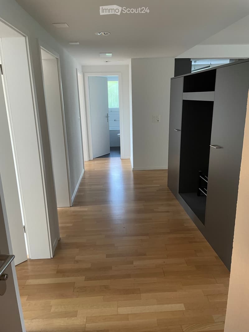 Empty hallway with wooden flooring, white walls, and recessed lighting. Features a built-in storage unit on the right and an open door leading to another room.