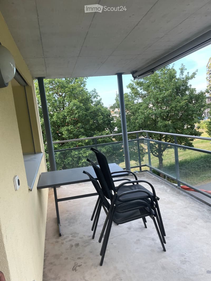 Balcony with a black table and several black chairs, overlooking a green area with trees.