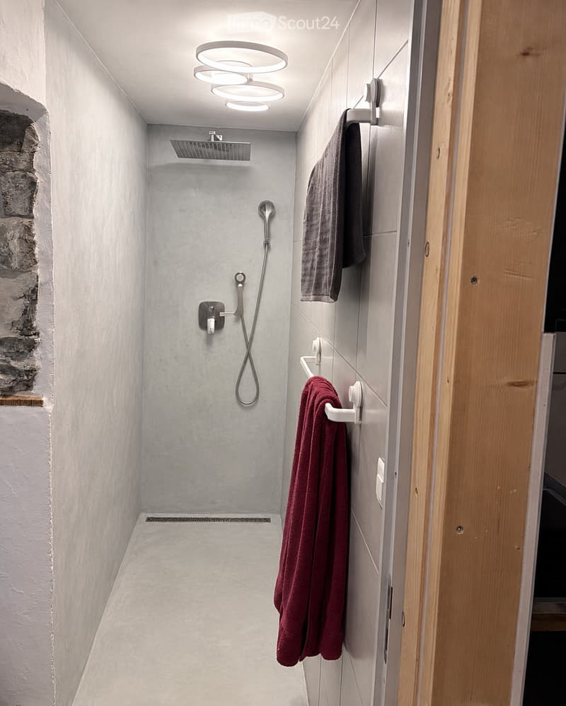 Modern, minimalist shower area with grey tiles, a handheld showerhead, and a towel rack holding a red towel.