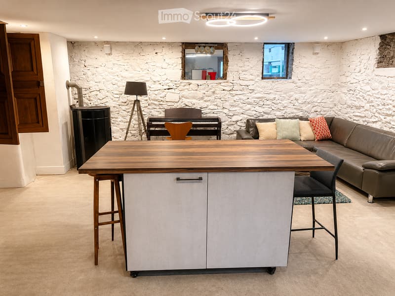 A modern, open-plan living space with a stone accent wall, a large wooden table with bar stools, a brown leather sofa, a piano, and a floor lamp.