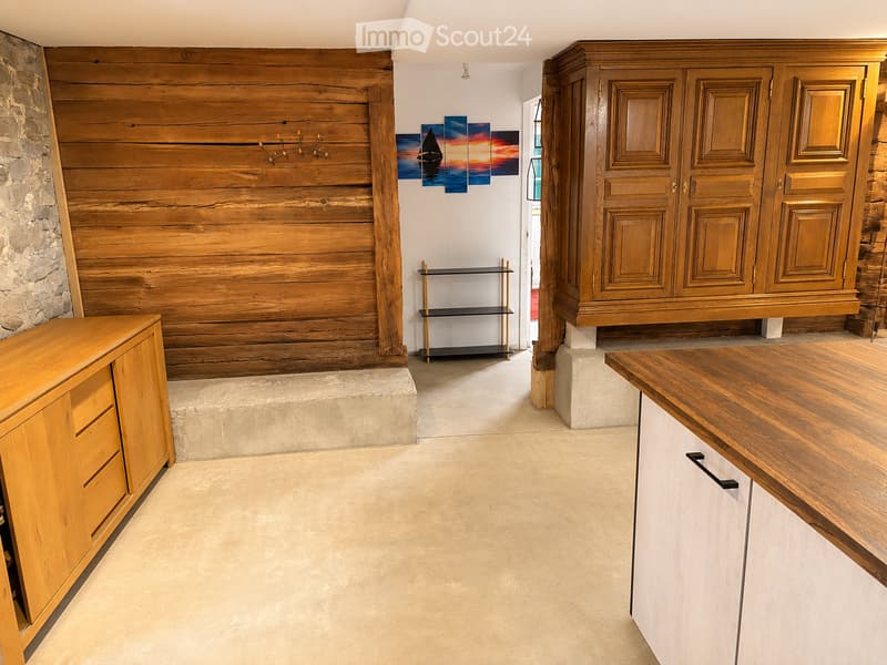 A spacious hallway with a wooden wall, stone wall, and a large wooden cabinet. There is a small black shelf and a wooden counter with white cabinets.