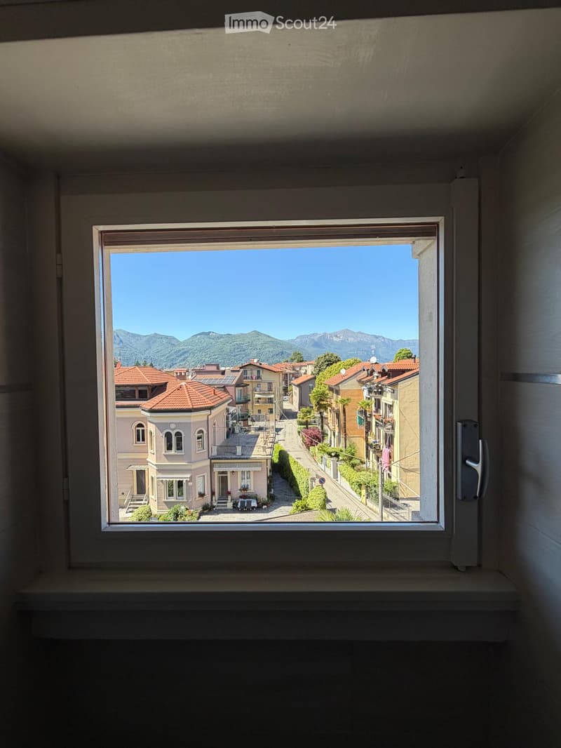An unfurnished room with a large window offering a scenic view of a town with red-tiled roofs, greenery, and distant mountains under a clear blue sky.