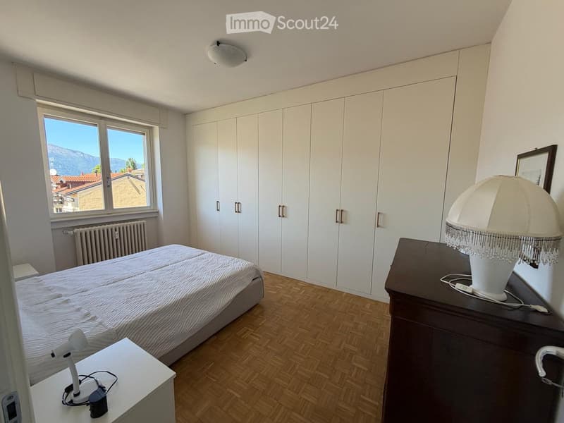 Modern bedroom with a large bed, nightstand with lamp, wardrobe, and a view of the city and mountains through a large window.