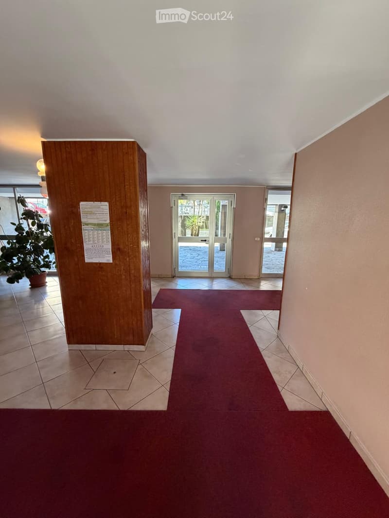 Empty hallway with tiled floor, red carpeted center, wooden pillar, and glass doors leading outside.