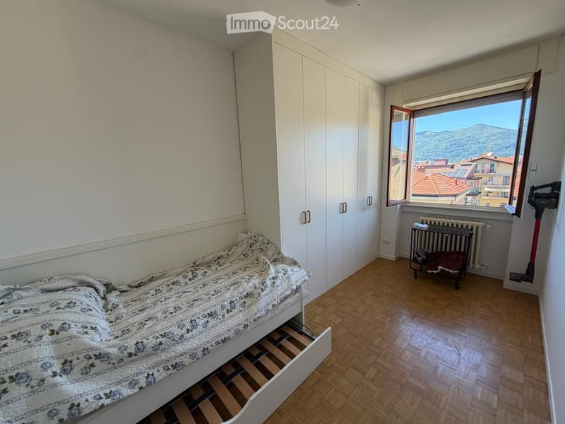 Bright bedroom with wooden flooring, a bed with patterned bedding, built-in white wardrobes, and a window offering a scenic view of the city and mountains.