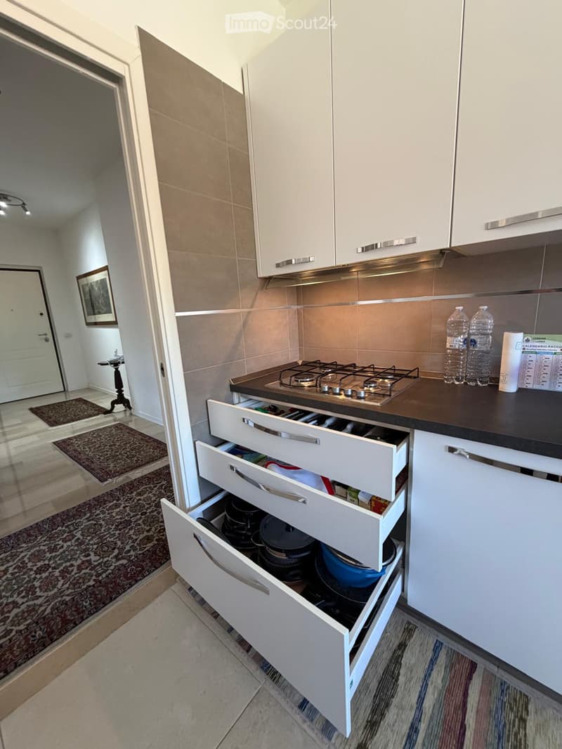 Modern kitchen with white cabinets, open drawers revealing pots and pans, a gas stove, and a countertop with water bottles.