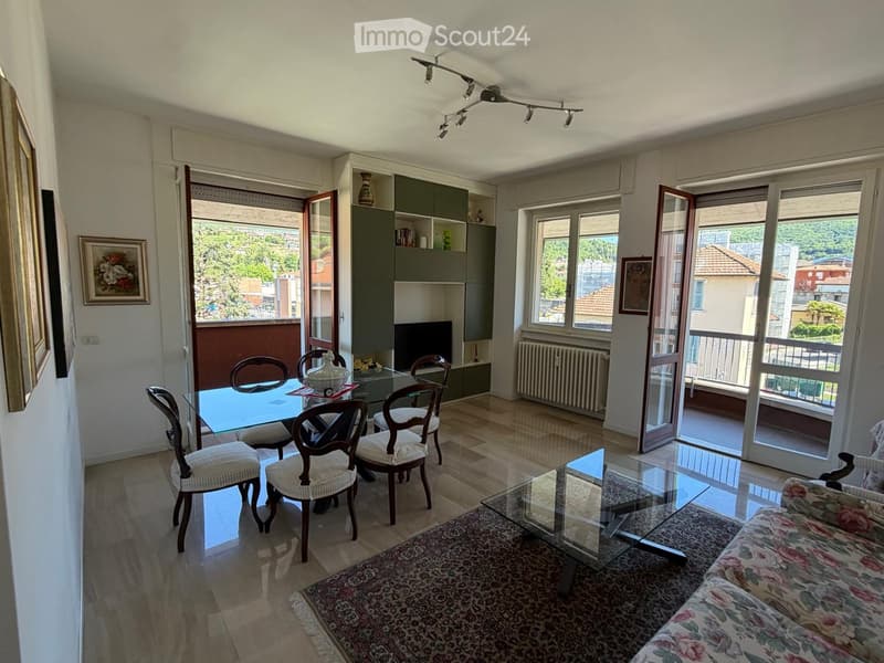 Bright living and dining area with wooden flooring, large windows, and sliding glass doors leading to a balcony. Features a sofa, coffee table, dining table with chairs, and a TV cabinet with a vase on top.