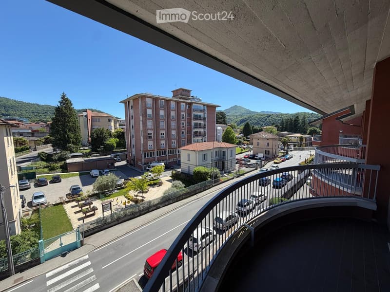 Balcony overlooking a street with parked cars, buildings, and a green area with trees and benches.