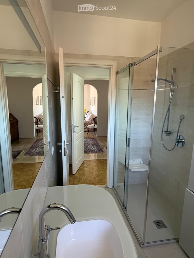 Modern bathroom with a freestanding bathtub, glass-enclosed shower, and toilet. The space features light-colored tiles and wooden flooring visible through an open doorway leading to an adjacent room.