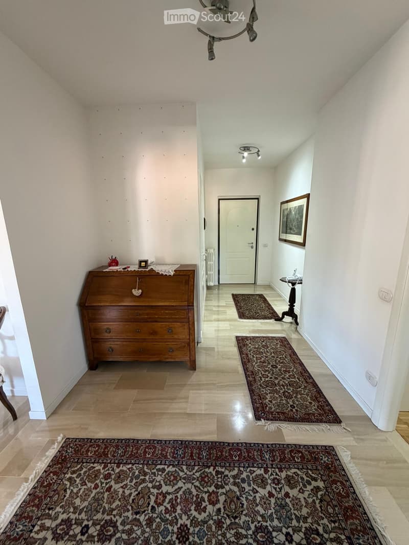 A well-lit hallway with wooden flooring, several patterned rugs, a wooden dresser with decorative items, and a framed picture on the wall.
