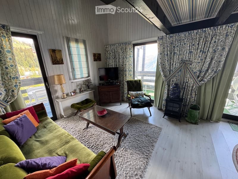 Cozy living room with wooden walls, large windows, green sofa, coffee table, armchair, ottoman, floor lamp, television, and cabinet.
