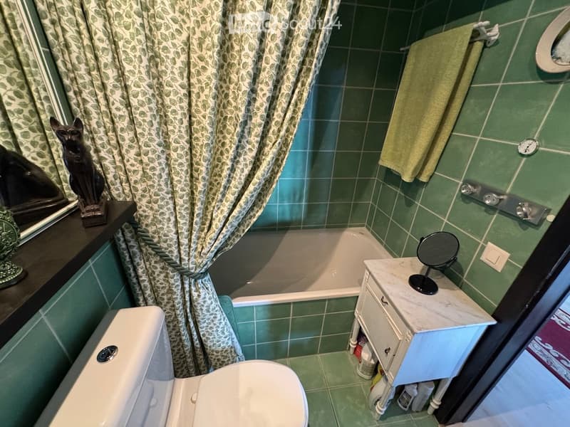 Bathroom with green tiled walls and floor, bathtub, toilet, small vanity with mirror, towel, and decorative statue on shelf.