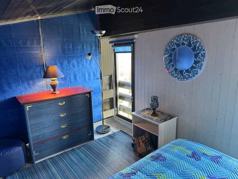 Cozy bedroom with a blue accent wall, a bed with a colorful quilt, a nightstand with a lamp, a drawer unit, a floor lamp, and a decorative mirror on the wall.