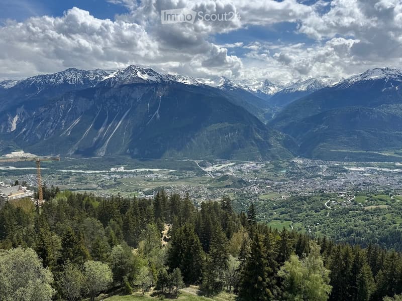 Panoramic view of a valley with snow-capped mountains, a town below, and a forested area in the foreground.