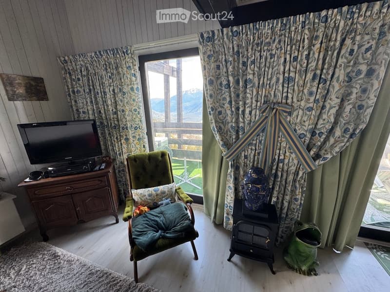 Cozy living room with a green armchair, television, wooden table, decorative vase, and a fireplace. Large windows offer a scenic mountain view.