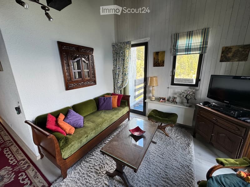 A cozy living room with a green sofa, colorful throw pillows, a glass coffee table, a wooden side table with a lamp, a television on a wooden stand, and a small green armchair.