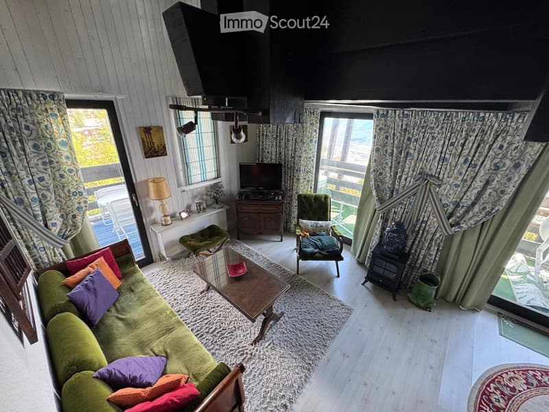 Cozy living room with a green sofa, colorful cushions, coffee table, side table, and a television. Large windows and a balcony are visible.