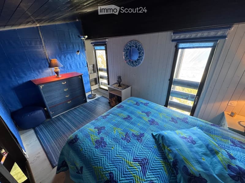 Cozy bedroom with a blue accent wall, a large window with blinds, a bed with a blue and green quilt, a nightstand with a lamp, a chest of drawers, and a small round mirror on the wall.