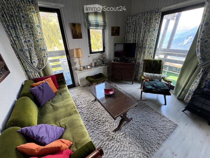 Cozy living room with a green sofa, coffee table, armchair, table lamp, television, and scenic mountain view through large windows.