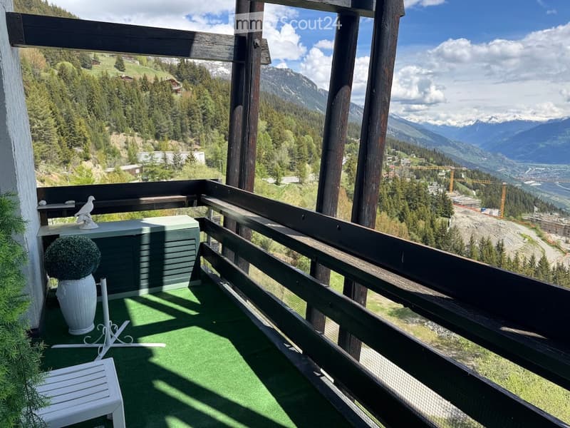 Balcony with green artificial turf, white chair, vase with plant, and decorative statue, overlooking a scenic mountain landscape.