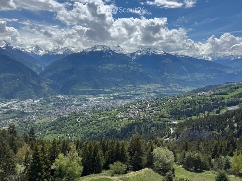 Panoramic view of a valley with snow-capped mountains, a town below, and dense forests covering the slopes.