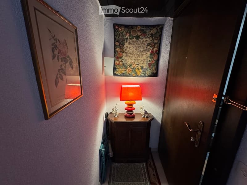 A narrow hallway with a small wooden table holding a lamp, framed artwork, and decorative items. The walls are painted white, and the floor is carpeted.