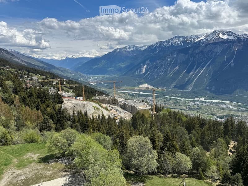 Panoramic view of a mountainous landscape with a construction site in the foreground, surrounded by dense forest and open fields.