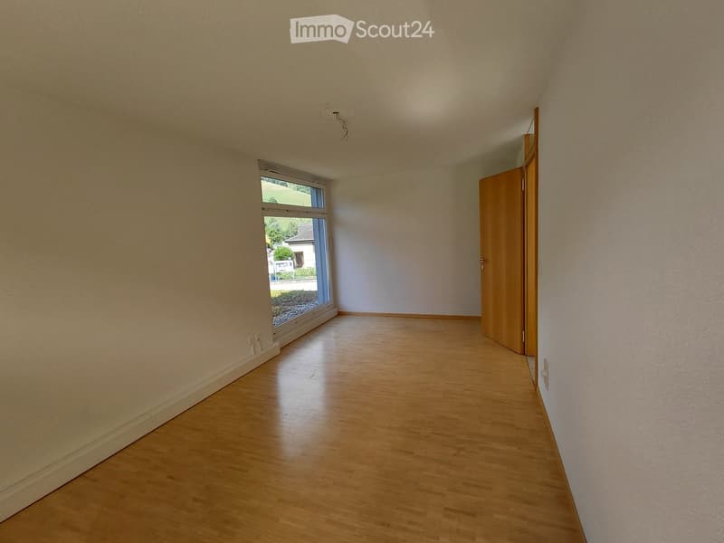 Empty room with wooden flooring, white walls, and a large window with a view of the outside. A wooden door is visible on the right side.