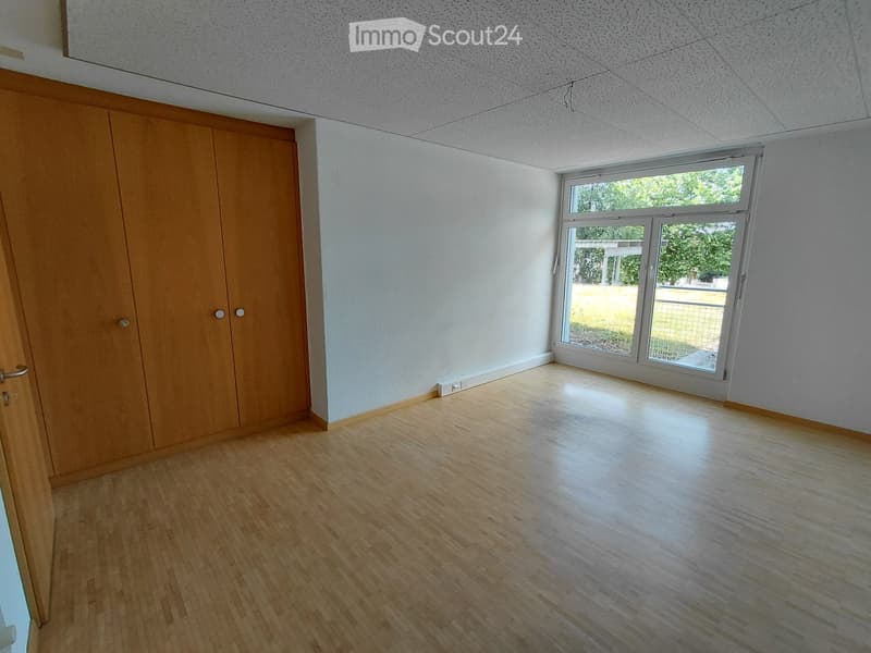 Empty room with wooden flooring, white walls, and a large window with a view of greenery outside. A wooden closet is visible on the left side.