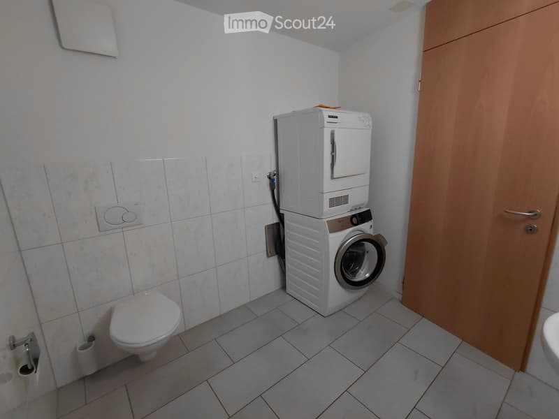 Modern bathroom with white tiled walls and floor, toilet, washing machine, and toilet paper holder.
