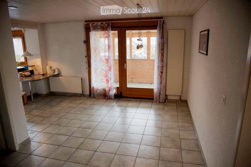 Bright hallway with tiled floor, wooden double doors leading outside, and a small kitchen area visible to the left.