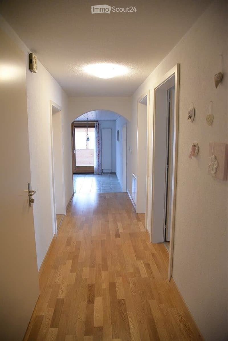 A long, narrow hallway with wooden flooring and white walls, featuring several doors and a recessed ceiling light. The hallway leads to an open area with a visible room beyond.