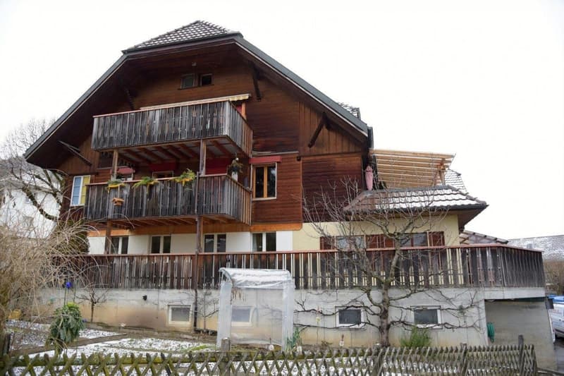 A two-story wooden house with a balcony, multiple windows, and a fenced yard.