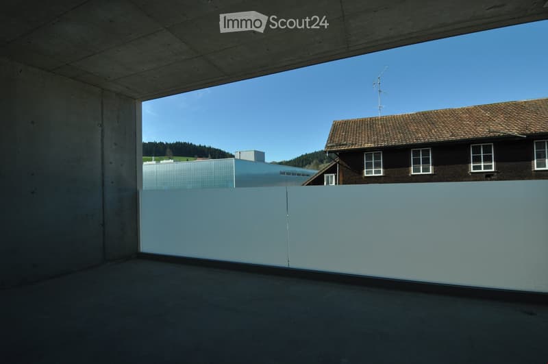 Minimalist balcony with concrete floor and walls, large glass panels, overlooking a landscape with a traditional house and modern building.