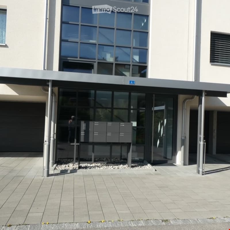Modern building entrance with a covered walkway, glass doors, and a row of mailboxes. The entrance features a clean, contemporary design with a paved area and a small garden bed with rocks.