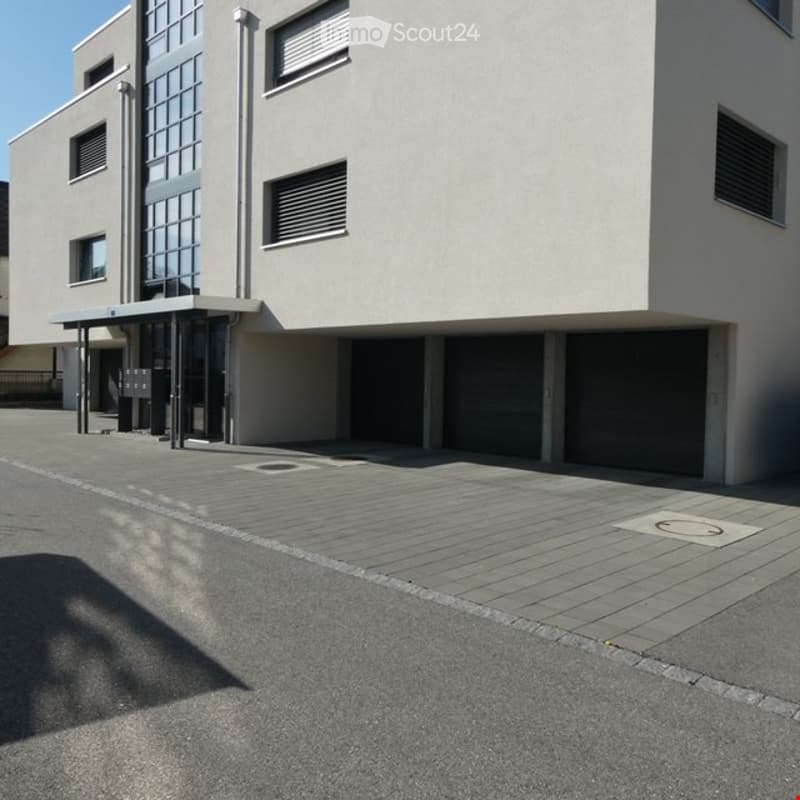 Modern multi-story building with a raised ground floor, large garage doors, and a paved entrance area.