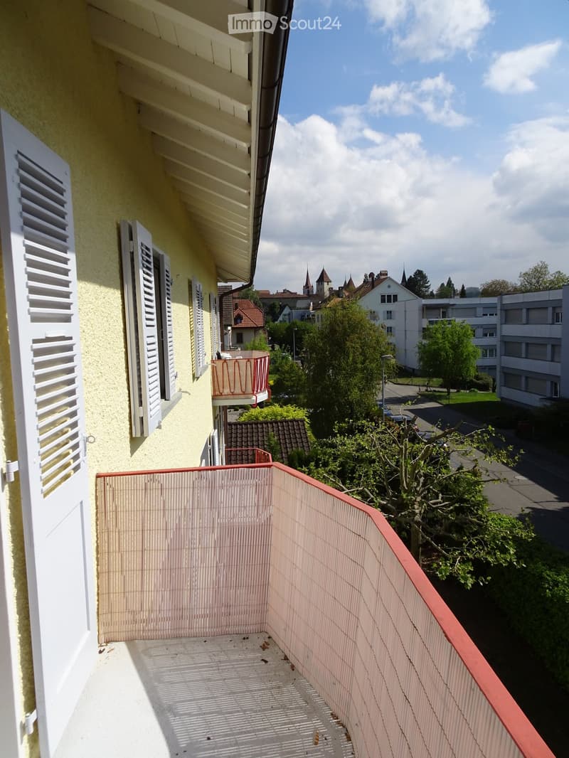 Balcony with beige tiled floor, red railing, overlooking a residential area with trees and buildings.