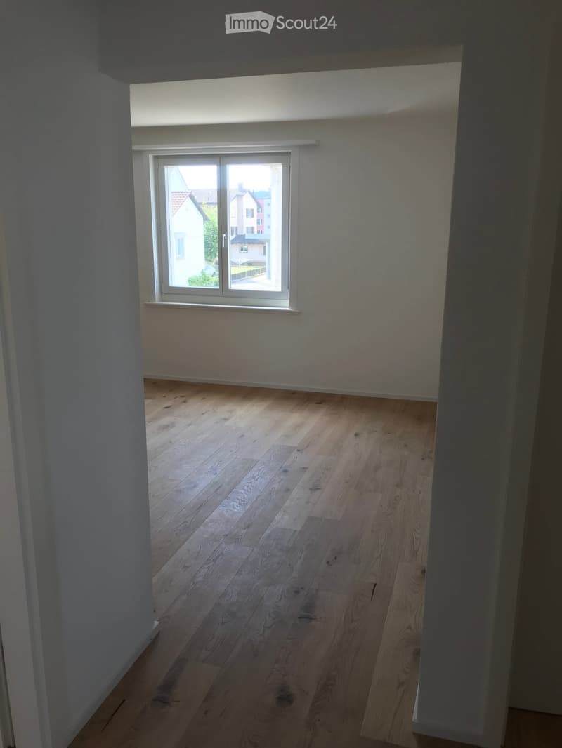 Empty room with wooden flooring and white walls, featuring a large window with white frame and view of neighboring houses.