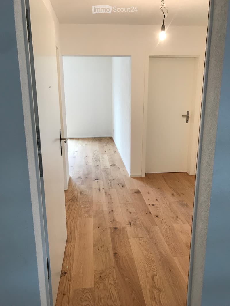 Empty hallway with light wooden flooring, white walls, and two doors, one open and one closed.