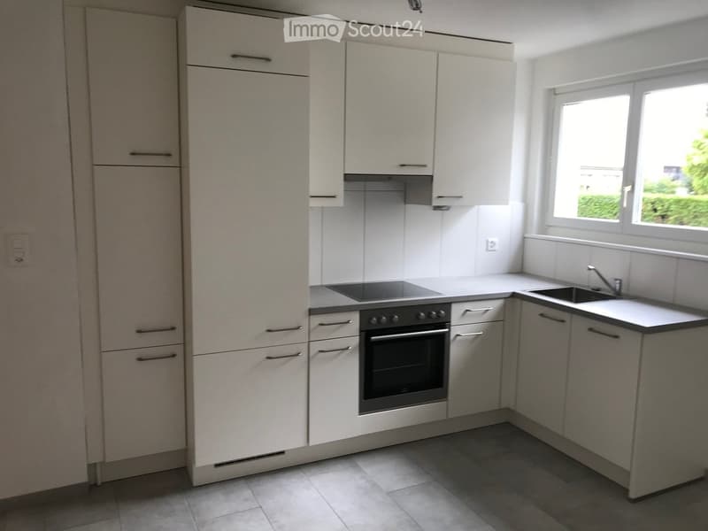Modern kitchen with white cabinets, stainless steel oven, sink, and countertop, featuring a window with a view of greenery.