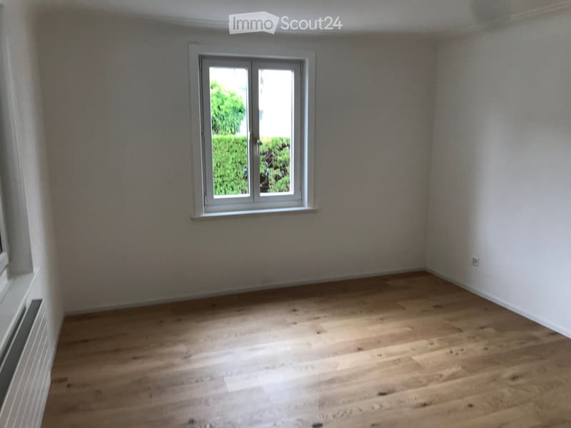 Empty room with wooden flooring, white walls, and a window showing greenery outside.