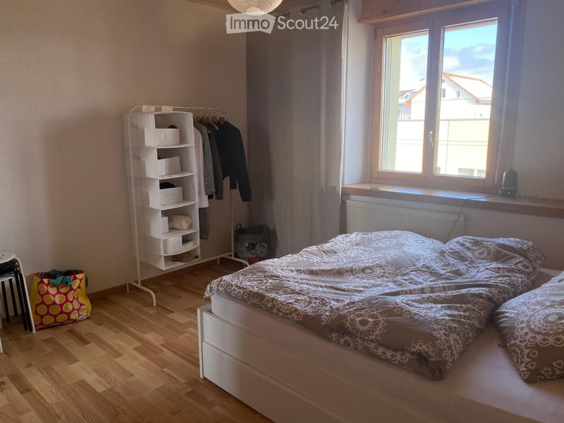 Bedroom with bed, white shelves, clothes hanging, window with curtains, wooden floor, bag, stool, and a basket.