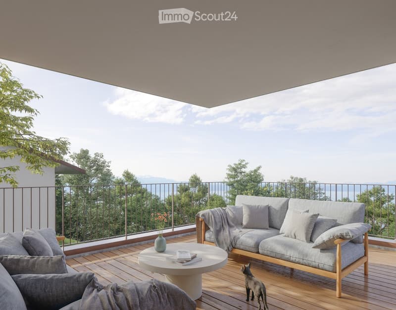 Balcony with wooden decking, grey sofa, coffee table, vase with flowers, books, and a small cat figurine. Offers scenic views of trees and mountains.