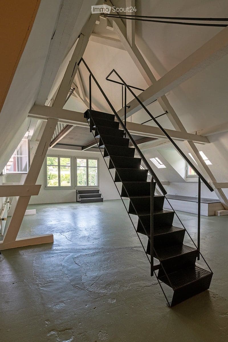 An empty attic space with exposed wooden beams, a modern staircase leading upwards, and large windows allowing natural light to enter.