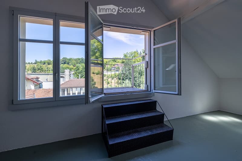 Empty room with large windows opening to a balcony, metal staircase leading up, and a view of greenery and rooftops.